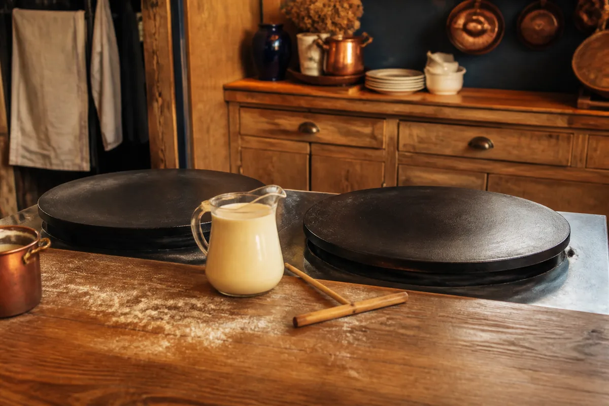 Galette de blé noir en cours de cuisson sur une plaque ronde, rozell posé sur le bord